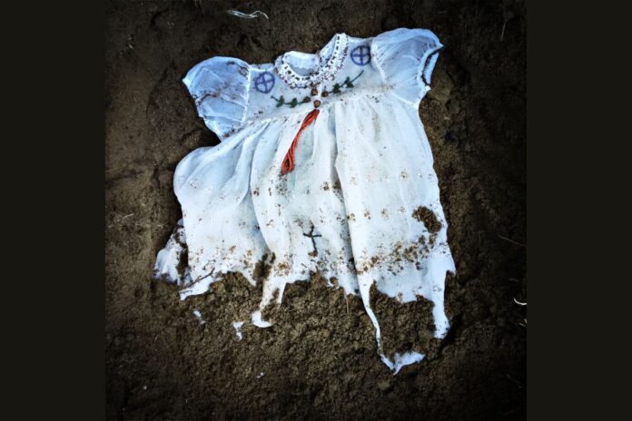 A Christian baptism gown with Indigenouss beadwork lies in the dirt.