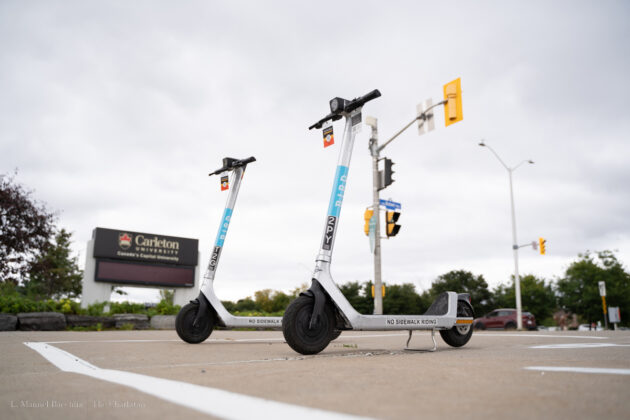 Two e-scooters parked with a Carleton University sign in the background.
