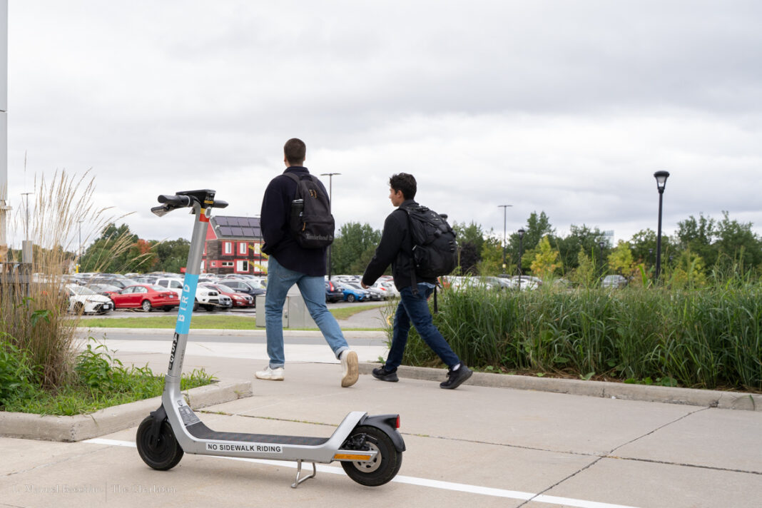 A white e-scooter pictures with two people walking in the background.