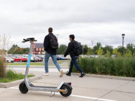 A white e-scooter pictures with two people walking in the background.