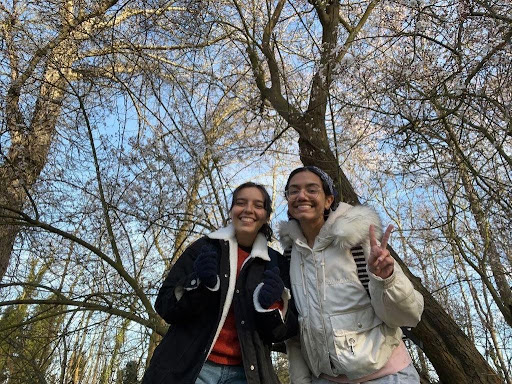 two girls have their arms around eachother and smile at the camera while standing in a forest
