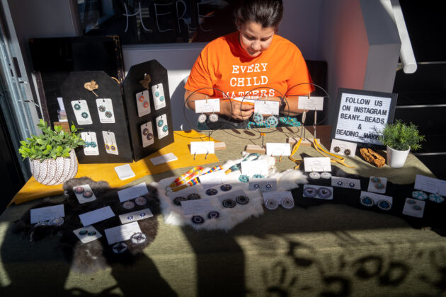 A woman wearing an orange shirt is sitting at a table with beads and other products for sale in front of her.