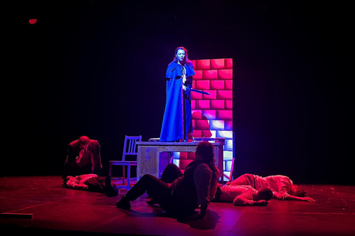 Lindsey Keene as Dracula stand up on a platform on stage with bodies laying on the stage around her.