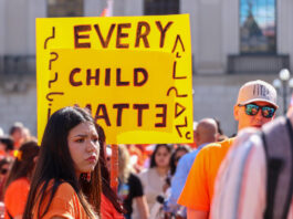 People wearing orange shirts standing below yellow signs that read "Every Child Matters."