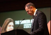 Carleton University launches award honouring Palestinian journalist Anton Abu Akleh speaks into a microphone in front on a screen displaying Shireen Abu Akleh's face.