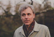 Q&A: Canadian music icon Joel Plaskett talks new album and his musical journey Man with white hair in a grey fall coat and wearing a red scarf looks away from the camera amid a background of winter-fall trees.