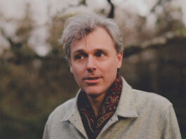 Man with white hair in a grey fall coat and wearing a red scarf looks away from the camera amid a background of winter-fall trees.