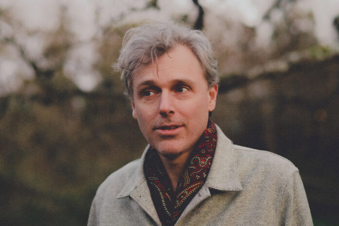 Man with white hair in a grey fall coat and wearing a red scarf looks away from the camera amid a background of winter-fall trees.