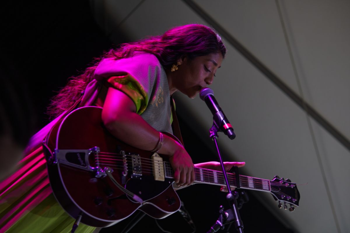 Women in a dress strums her guitar and sings on stage in a canted angle. 