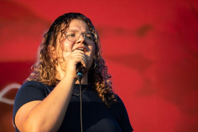 Woman with curly hair and wearing a black top sings in front of a red backdrop.