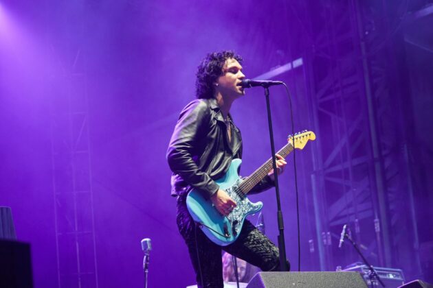 Man in all black outfit with light blue guitar strums his guitar and sings on stage.