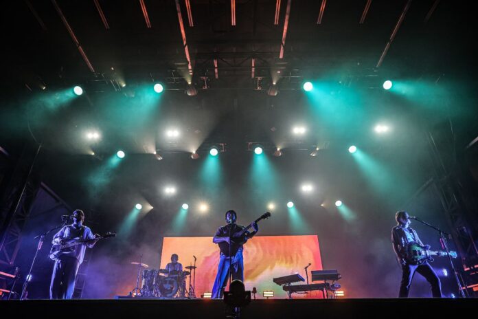 Green and purple strobe lights flash as a 2 guitarist, a drummer, and keyboardist are on stage.