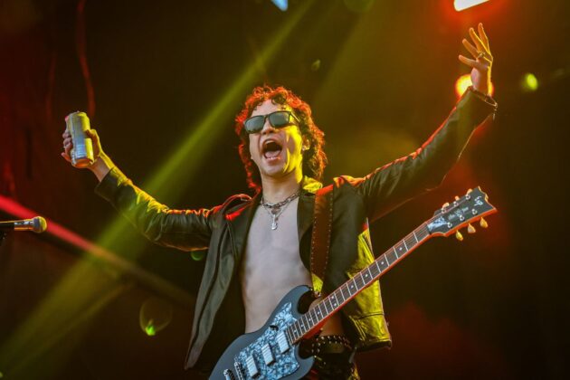 Man in all black with sunglasses on hold a drink can in one hand and has his hands up in the air. He is yelling on stage.