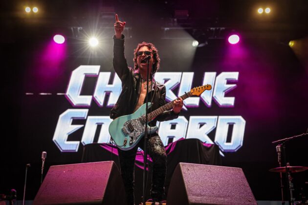 Man in all black outfit with light blue guitar strums his guitar and sings on stage.