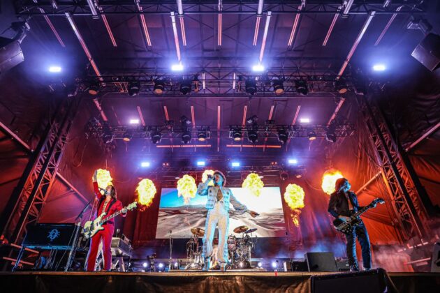 With flames going off at the side of the stage, the lead singer in an all white bedazzled outfit sings with his band on stage.