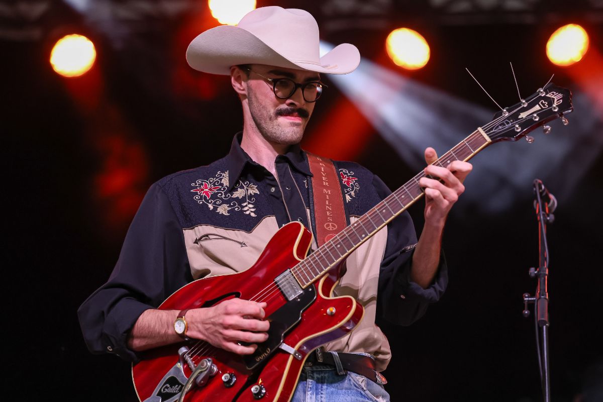 Man in a white cowboy hat with a red guitar passionately plays the guitar on stage. 