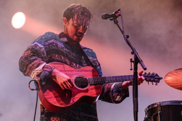 Man in a blue poncho with a red guitar strums his guitar in front of a mic stand on stage.