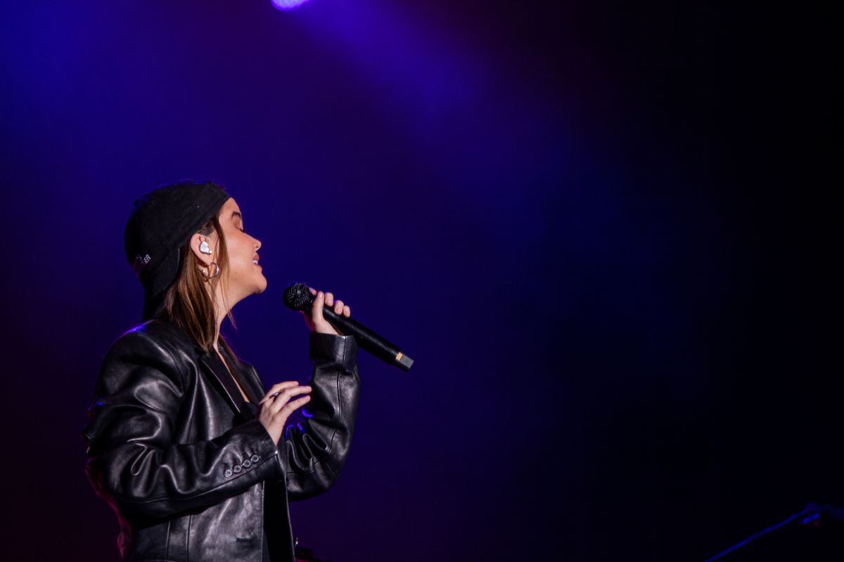 Woman in all black and a black cap sings on stage with a black mic amid a blue backdrop.