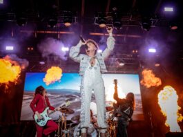 With flames going off at the side of the stage, the lead singer in an all-white bedazzled outfit sings with his band on stage.