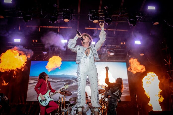 With flames going off at the side of the stage, the lead singer in an all-white bedazzled outfit sings with his band on stage.