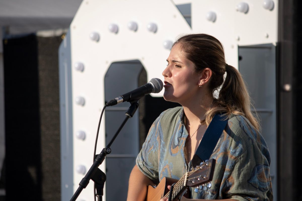 Women with a ponytail and a grey shirt sings in front of a mic with a guitar. 