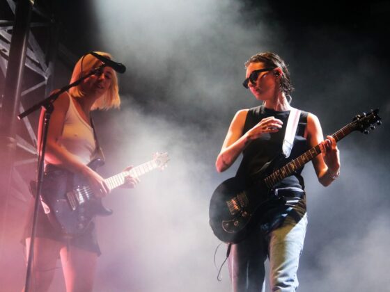 Two guitarists strum their guitar next to one mic on stage.