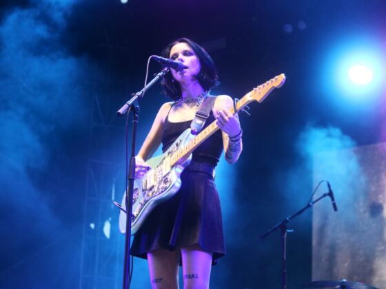 Women wearing black mini dress strums a white guitar on stage with moody blue lighting.