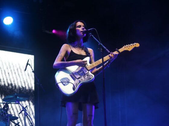 Women wearing black mini dress strums a white guitar on stage with moody blue lighting.