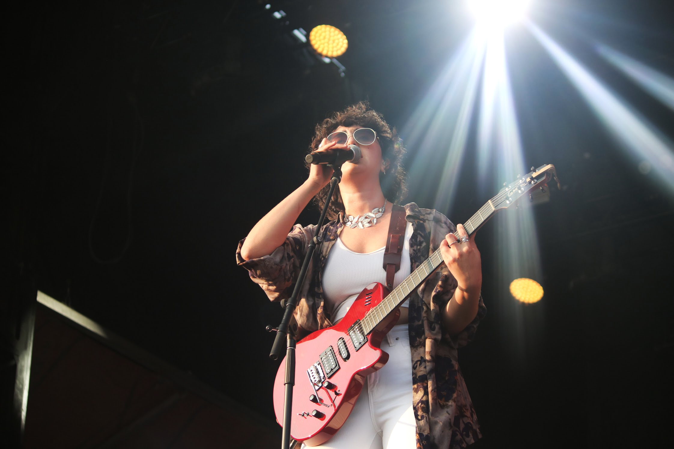 Singer with a red guitar and sunglasses hold the mic and sings on stage. 