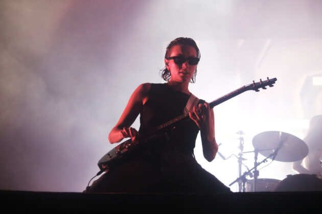 Women wearing all black with black sunglasses kneels on stage and strums her guitar hard amidst red lighting.