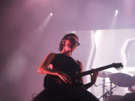 Women wearing all black with black sunglasses kneels on stage and strums her guitar hard amidst red lighting.