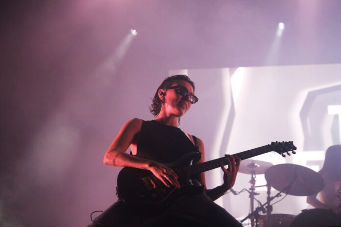 Women wearing all black with black sunglasses kneels on stage and strums her guitar hard amidst red lighting.