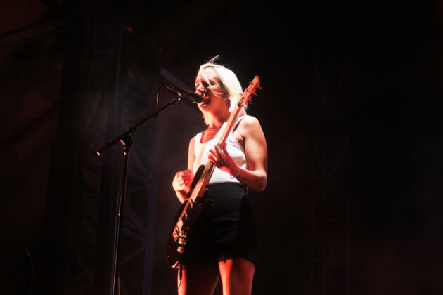 Blonde women wearing a white tank top and black mini skirt strums her guitar in front of a mic stand.