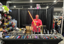Ottawa Comiccon embraces vendors, hobbyists and Hobbit mania Man in red t-shirt poses with a red lightsaber in his booth at Comiccon. On the table in front of him are other light sabers of various colours that he is selling.