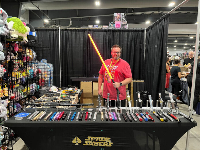 Man in red t-shirt poses with a red lightsaber in his booth at Comiccon. On the table in front of him are other light sabers of various colours that he is selling.