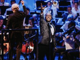 A Black man in a suit sings with his right hand extended in the air in passion. In the back, the NAC orchestra is seated and playing along.