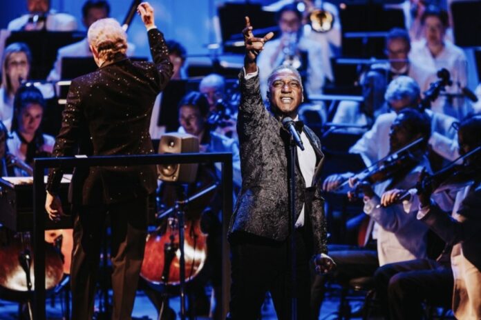 A Black man in a suit sings with his right hand extended in the air in passion. In the back, the NAC orchestra is seated and playing along.