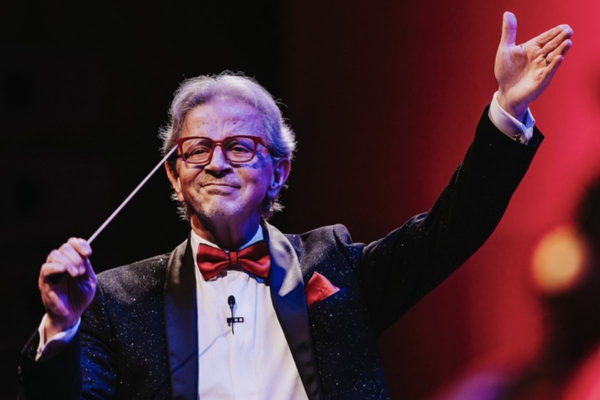 A man wearing a black suit with a red bowtie closes his eyes as he waves the conductor stick in front of his face with passion. 