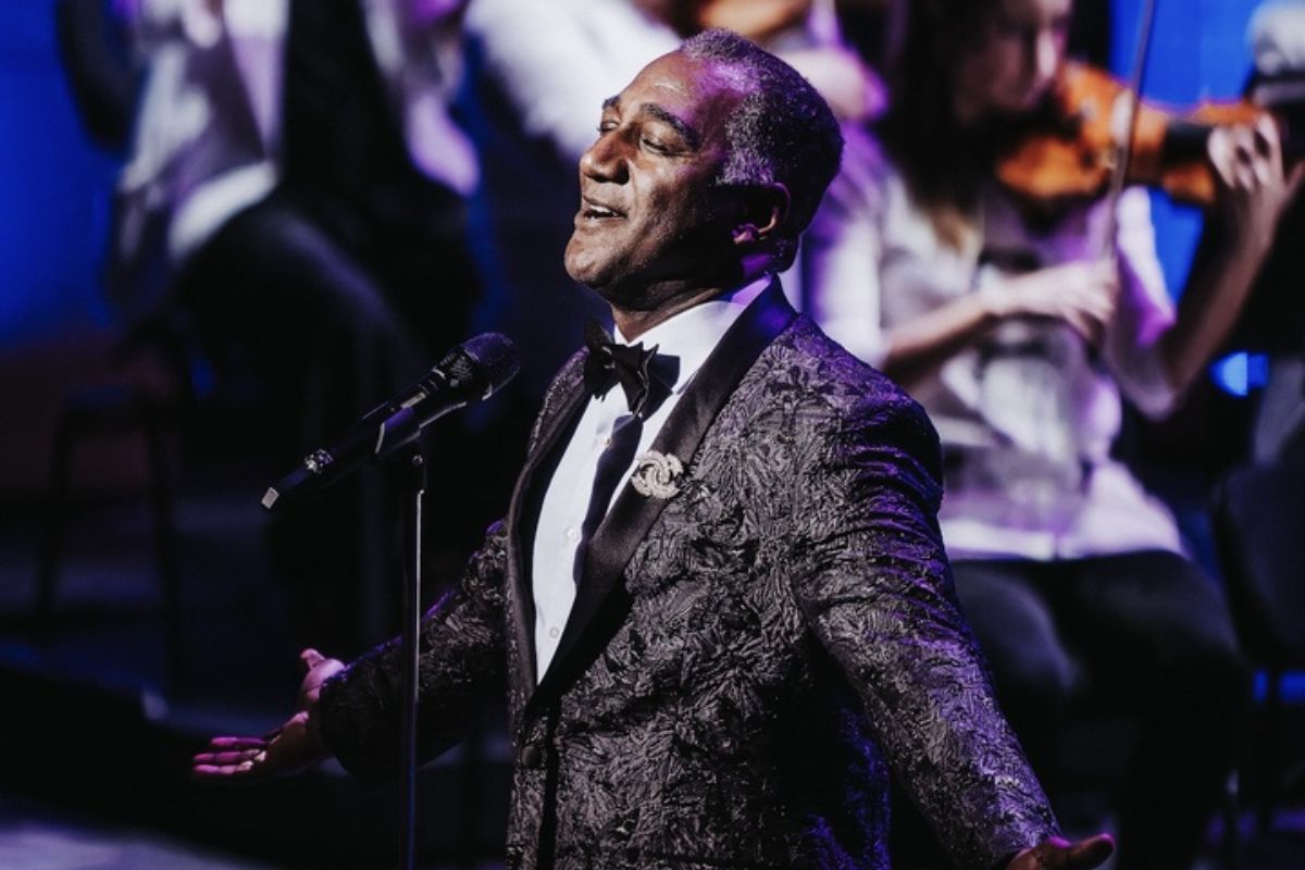 A Black man wearing a black suit with some details on it closes his eyes and sings in front of a mic with the NAC orchestra behind him. 