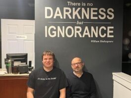 Underneath a sign that reads "There is no darkness but ignorance," two men sit wearing black shirts. The one on the left is a blind server, while the one on the right is the owner of the restuarant.