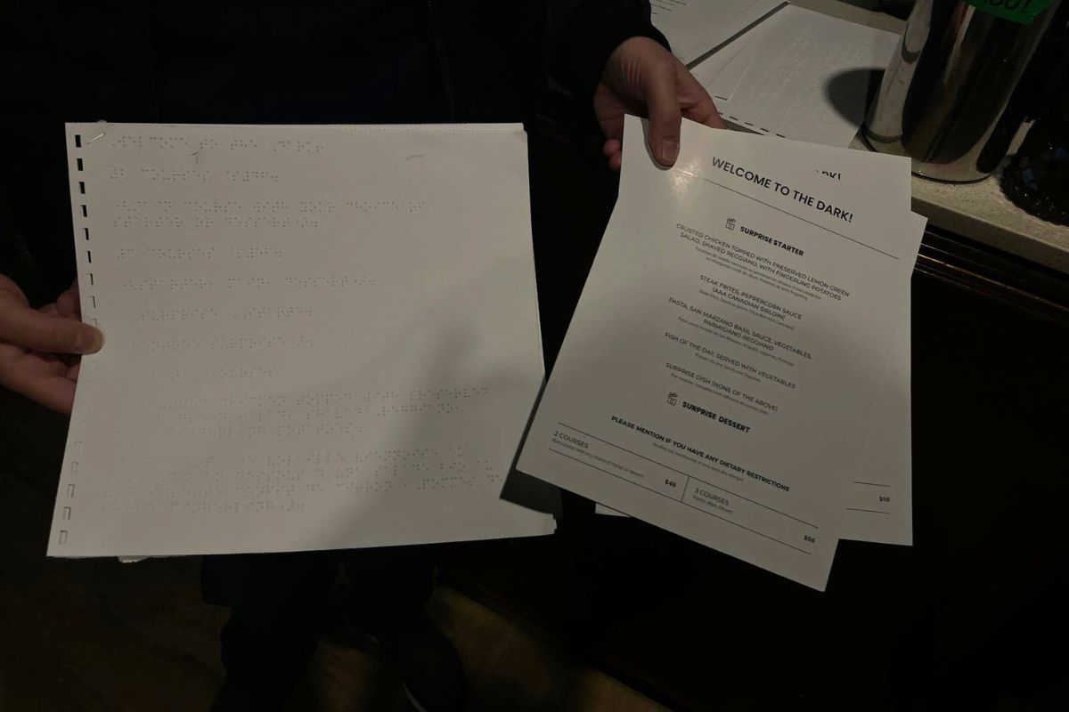 A hand holds out two menus, on the right is a white menu in braille with the one on the left is a white menu with black writing on it. 