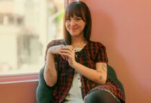 Q&A: Gracie-May Saint returns to traditional hand-poked tattoo methods Against a pink background, a women with tattoos wearing a red plaid shirt smiles at the camera.