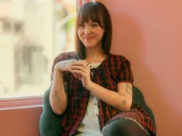 Against a pink background, a women with tattoos wearing a red plaid shirt smiles at the camera.