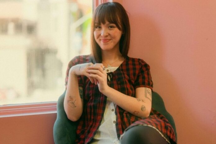 Against a pink background, a women with tattoos wearing a red plaid shirt smiles at the camera.