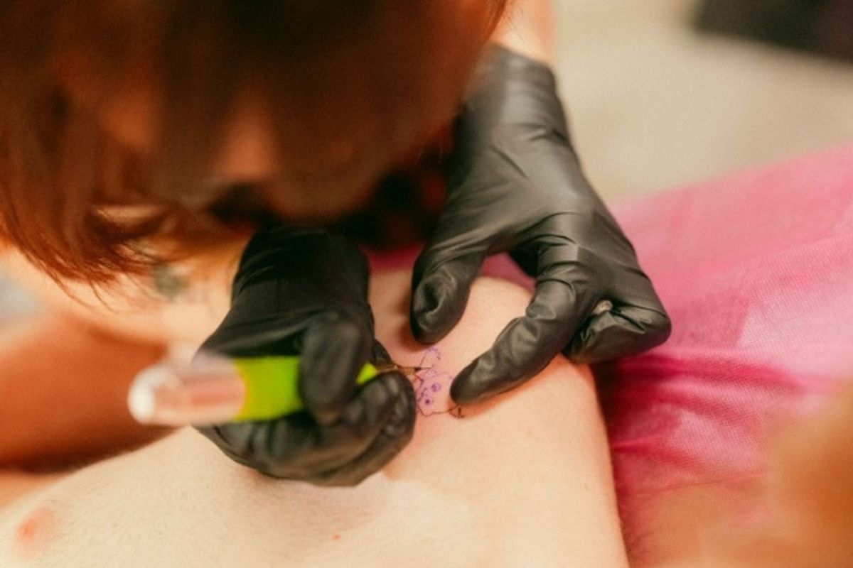 Close up shot of a white arm, and a women holding a tattoo needle making a lil angel.