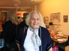 An elderly woman wearing a white blouse with a blue floral scarf smiles for the camera.