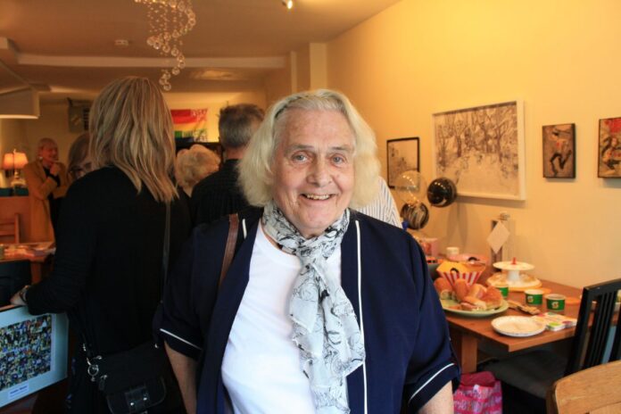 An elderly woman wearing a white blouse with a blue floral scarf smiles for the camera.