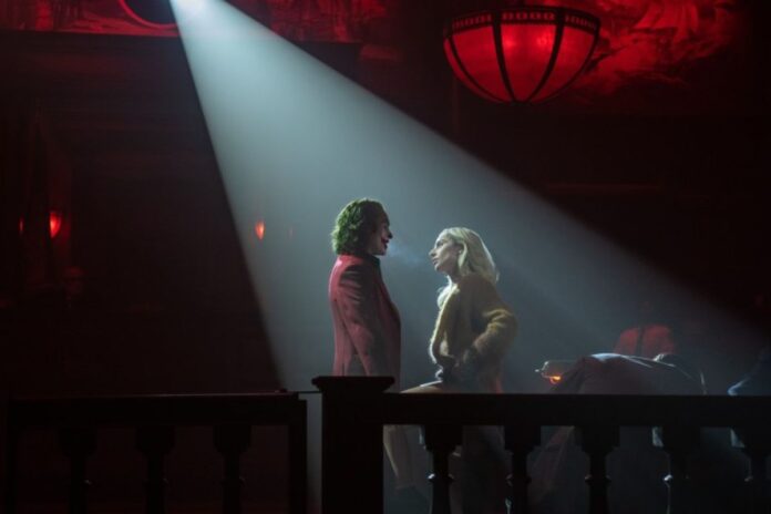 With a white spotlight on them, Joker and Harley Quinn talk in a dark restaurant