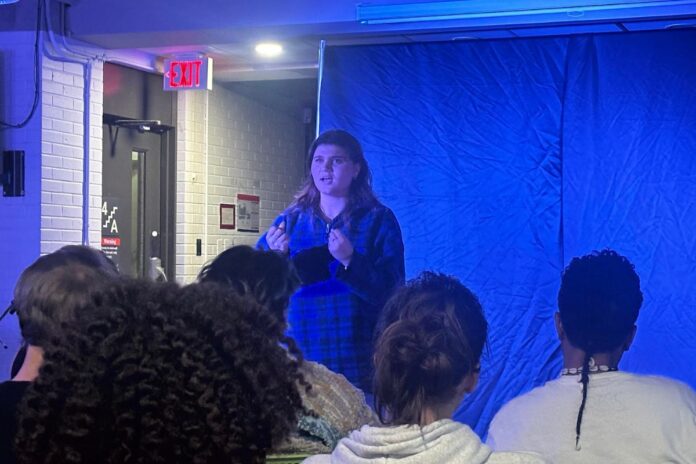 Amid a blue background and a crowd watching, a girl in a blue bottom down shirt acts out a scene from The Edge of Seventeen.