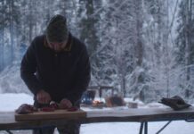 Review: ‘My Indian Name’ tells a poignant and powerful story Admid a snowy background of frozen trees, a man in a hat and black jacket cut meat at an outside table.